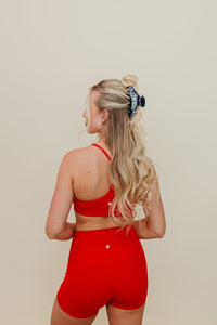 Woman wearing a red sports bra and shorts wearing black and white gym girl claw on a beige background