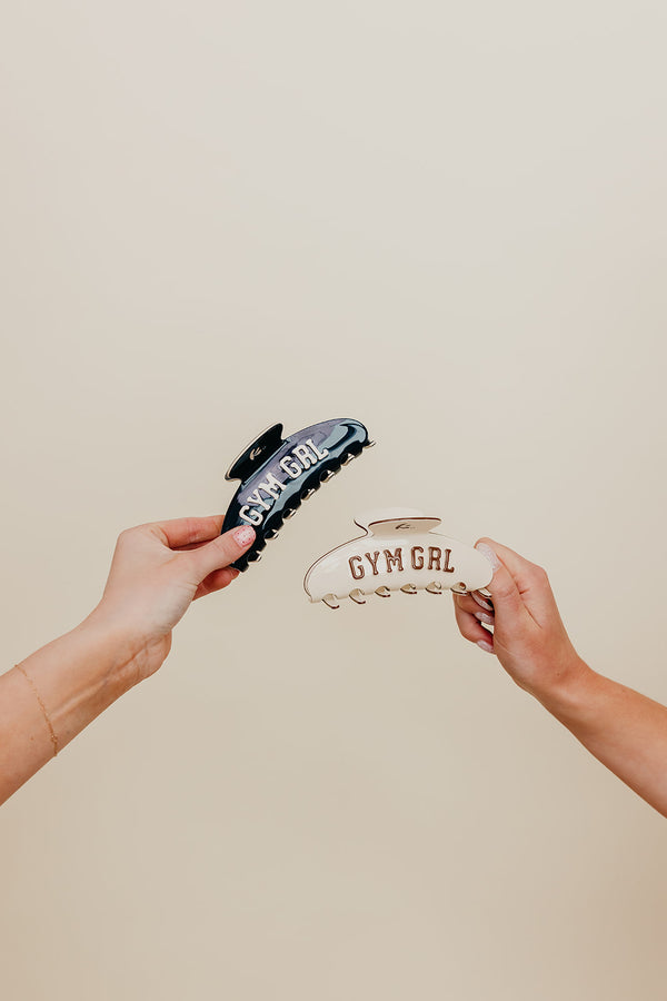 Two hands holding navy and cream claw with 'Gym Girl' text against a beige background