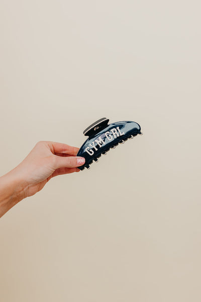 Hand holding a black hair clip with 'Gym Girl' text on a beige background