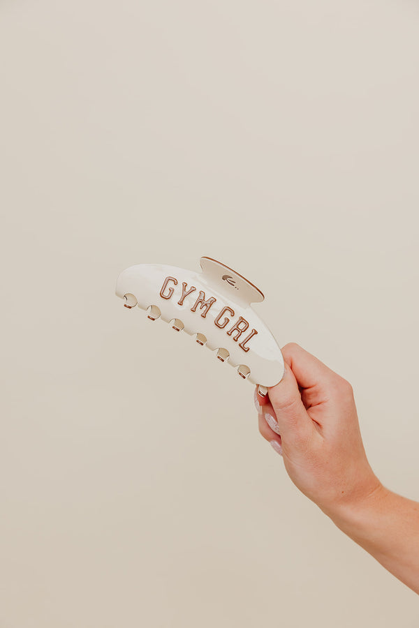 Hand holding a cream and brown claw with 'GYM GIRL' text on a beige background