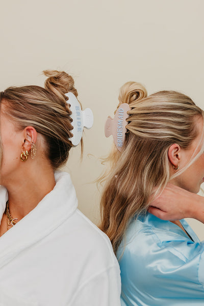 Two women with styled hair, one in a white robe and the other in a blue robe, wearing bride and bridesmaid claw against a neutral background.