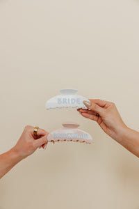 Two hands holding hair clips with 'Bride' and 'Bridesmaid' text on a beige background