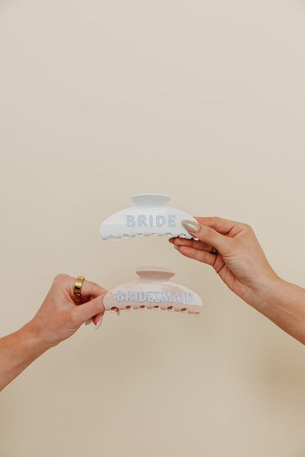 Two hands holding hair clips with 'Bride' and 'Bridesmaid' text on a beige background