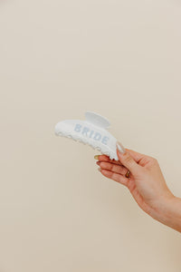 Hand hold white and blue claw with 'BRIDE' text held by a hand against a beige background