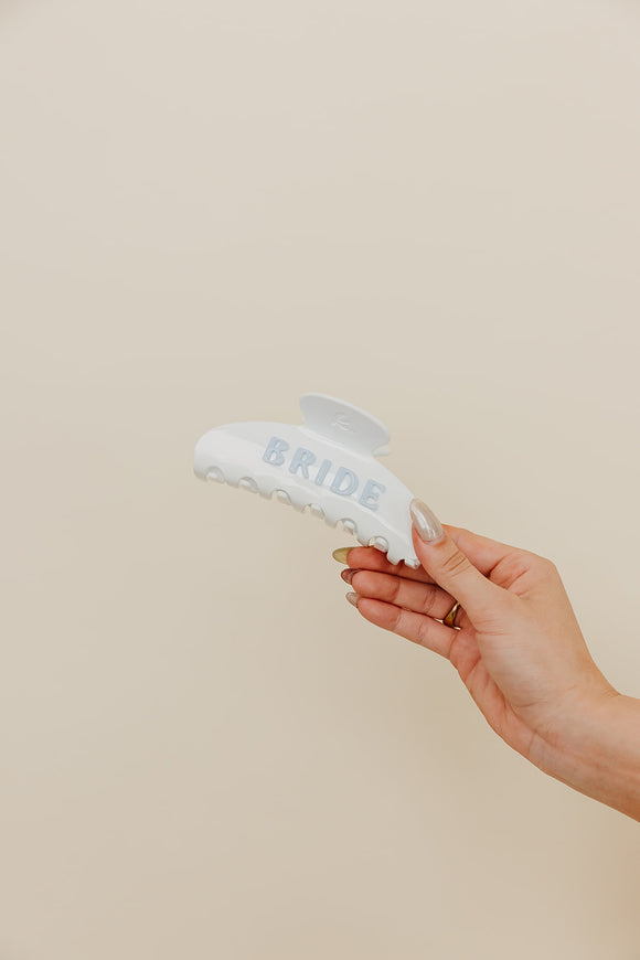 Hand hold white and blue claw with 'BRIDE' text held by a hand against a beige background